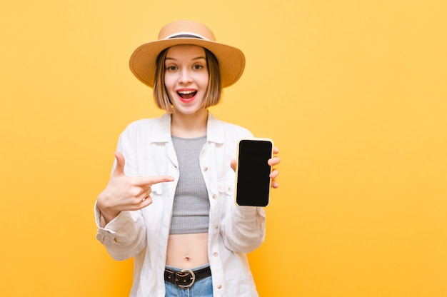 Turista de garota com roupas de chapéu e verão mostra smartphone com tela preta e dedo na tela, olha para a câmera com cara feliz