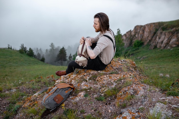 Turista de hipster brutal vestido em estilo vintage senta-se com mochila e manta em pedra na natureza