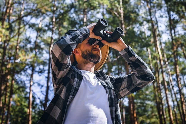 Turista de homem com um chapéu e uma camisa xadrez parece através de binóculos na floresta.