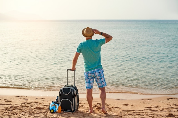 Turista de homem em roupas de verão com uma mala na mão, olhando para o mar na praia