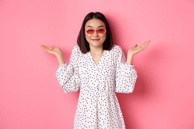 Turista de linda mulher asiática sorrindo para a câmera, encolhendo os ombros sem noção, não sei, usando óculos escuros da moda e vestido branco, em pé contra rosa.