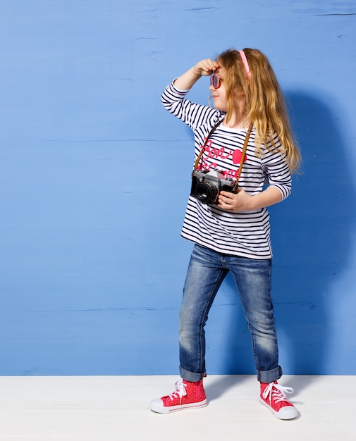 Foto turista de menina criança feliz com câmera retro na parede azul.