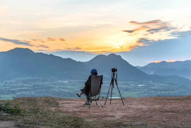 Turista de mulher asiática sentada em uma cadeira na montanha ao pôr do sol com uma câmera ao lado