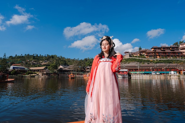 Turista de mulher asiática vestindo vermelho hanfu antigo vestido étnico tradicional chinês em pé no barco de madeira no reservatório da vila tailandesa de ban rak, província de mae hong son, tailândia