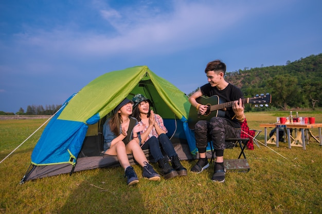 Turista de um grupo de amigos asiáticos bebendo e tocando violão junto com a felicidade no verão enquanto acampam perto do lago ao pôr do sol