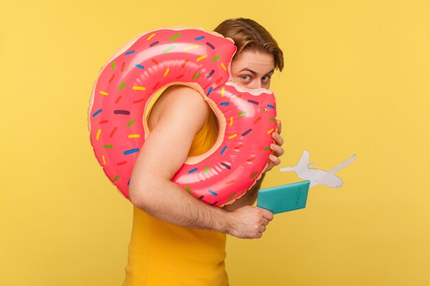 Un turista divertido con camiseta escondiéndose detrás de un anillo de goma de donut rosa, sosteniendo un documento de pasaporte y una maqueta de avión, mirando a la cámara tímidamente inseguro. tiro de estudio interior aislado sobre fondo amarillo