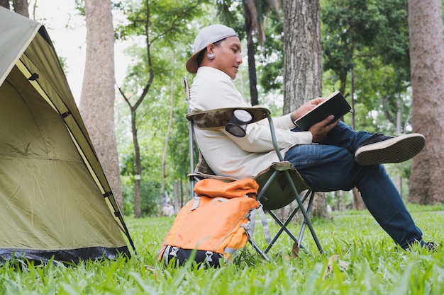 Turista do jovem sentado na cadeira e lendo o livro na frente da barraca no parque de campismo na floresta.