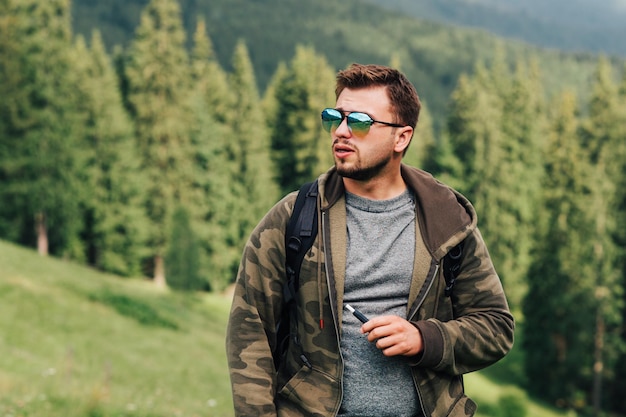 Turista do sexo masculino em uma expedição de montanha fez uma pausa de fumar um cigarro em pé em um prado contra o pano de fundo de uma paisagem natural irreal