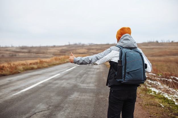 Turista do sexo masculino pega um carro na beira de uma estrada vazia de inverno em uma área suburbana com uma mochila. Um homem com chapéu laranja em uma rodovia.