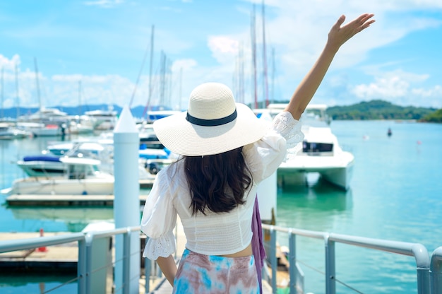 Un turista emocionado con sombrero blanco disfrutando y de pie en el muelle con yates de lujo durante el verano