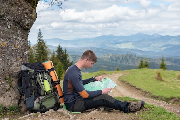 Turista está estudiando un mapa de la zona sentada debajo de un árbol junto a la carretera