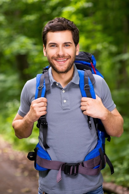 Turista feliz. Apuesto joven llevando mochila y mirando a cámara con una sonrisa mientras está de pie en la naturaleza