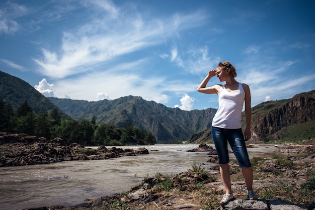 Turista feliz en una camiseta blanca y pantalones cortos de mezclilla se encuentra en la orilla del río de montaña en un día soleado.