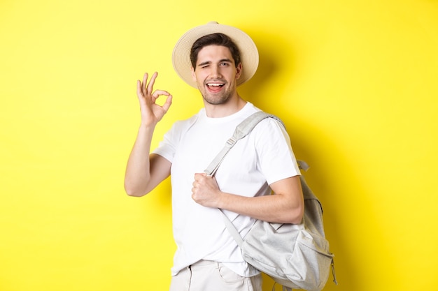 Turista feliz saindo de férias, segurando uma mochila e mostrando a placa de ok sorrindo satisfeito, em pé contra a parede amarela