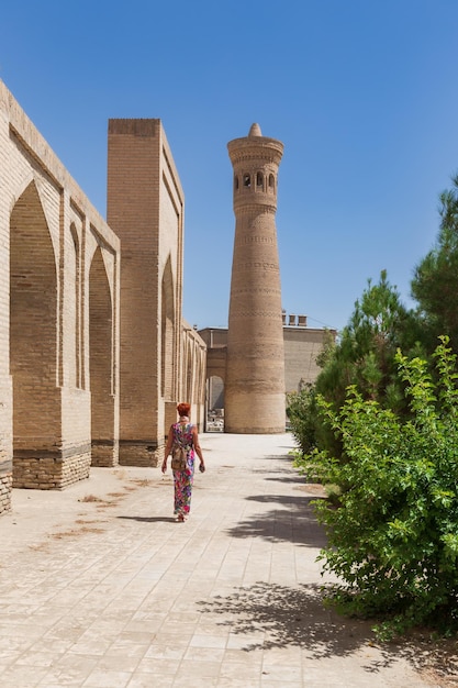 Turista feminina caminha ao longo da antiga parede da madrassa em direção ao minarete de tijolos na antiga cidade de Bukhara Uzbequistão