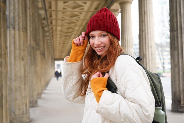 Turista feminina com cabelo comprido ruivo segura mochila com garrafa térmica se volta para a câmera com um sorriso feliz