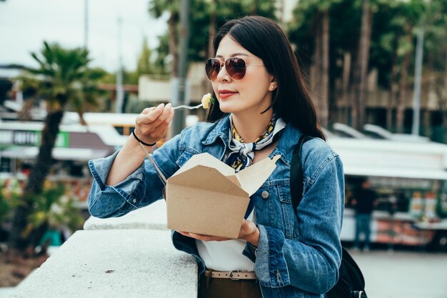 turista feminina na moda retrato está de pé contra o parapeito de pedra e almoçando. mulher feliz usa óculos de sol está desfrutando de batata assada e olhando a bela vista distante. momentos reais