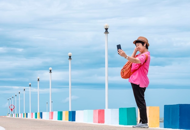 Turista feminina usando smartphone para tirar selfie em pé na ponte no ponto de vista do mar