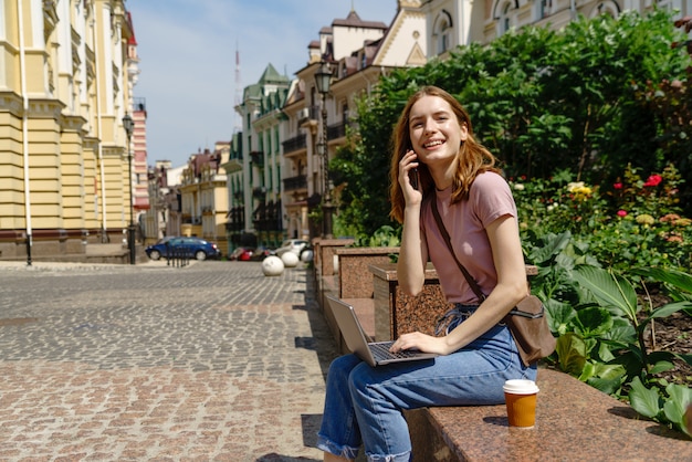 Turista hermosa joven con café para llevar en el centro de la ciudad usando la computadora portátil.