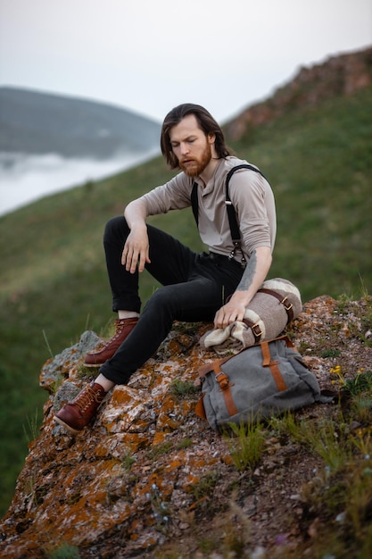 Turista hipster brutal vestido con estilo vintage se sienta con mochila y tela escocesa en piedra en la naturaleza