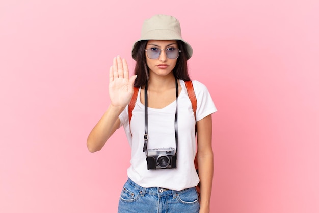 Turista hispânica bonita olhando a sério, mostrando a palma da mão aberta, fazendo gesto de pare com uma câmera fotográfica e um chapéu