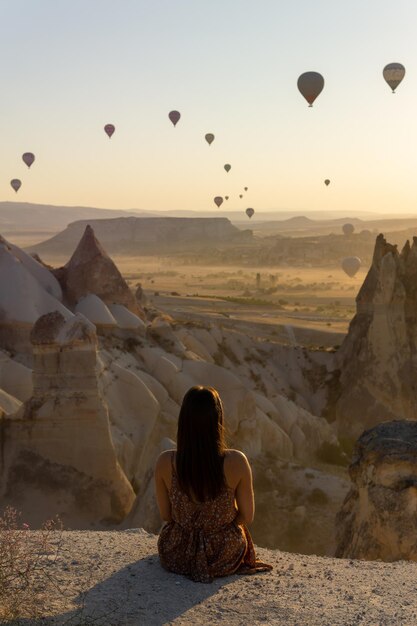 Turista irreconocible sentado en primer plano disfrutando de las vistas del valle del amor y globos aerostáticos volando al amanecer en Capadocia Turquía