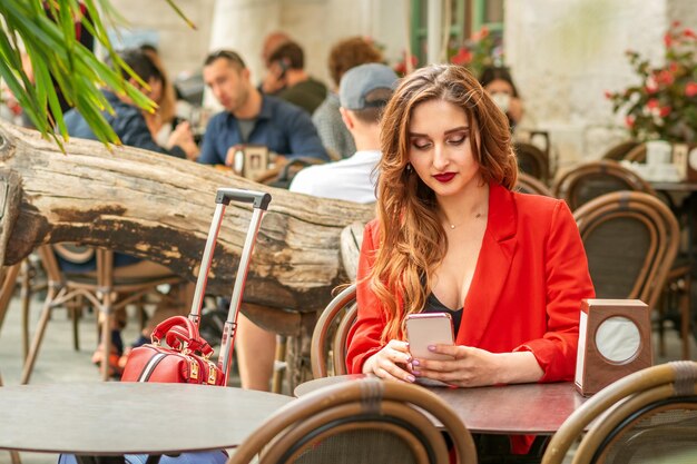 Turista, jovem branca, branca, com uma jaqueta vermelha, olhando para o smartphone sentado à mesa