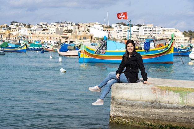 Turista jovem sentada e ao fundo o mar cheio de barcos coloridos em Marsaxlokk