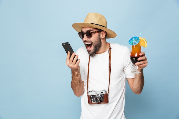 Foto turista joven emocionado guapo posando con la cámara mediante teléfono móvil bebiendo cócteles aislado sobre la pared azul.