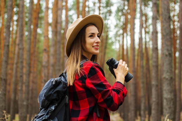 Turista joven con mochila, sombrero y camisa a cuadros roja con binoculares en el bosque.