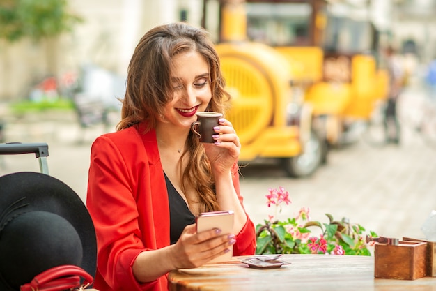 Turista joven mujer caucásica en chaqueta roja con taza de café en la mesa de café al aire libre.