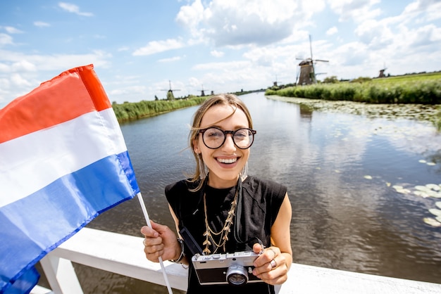 Turista joven de pie con la bandera holandesa en el hermoso paisaje de fondo con viejos molinos de viento en Holanda
