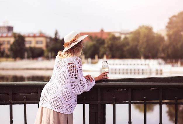 Turista joven de pie con una copa de vino blanco en el puente en el río de la ciudad europea al fondo