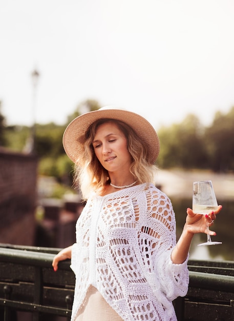 Turista joven de pie con una copa de vino blanco en el puente en el río de la ciudad europea al fondo