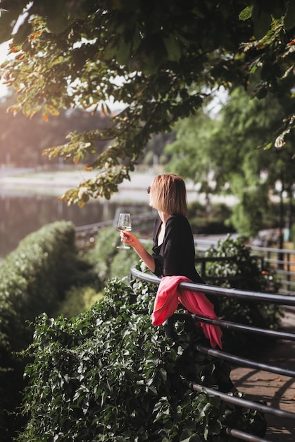 Turista joven de pie con una copa de vino blanco en el puente en el río de la ciudad europea al fondo