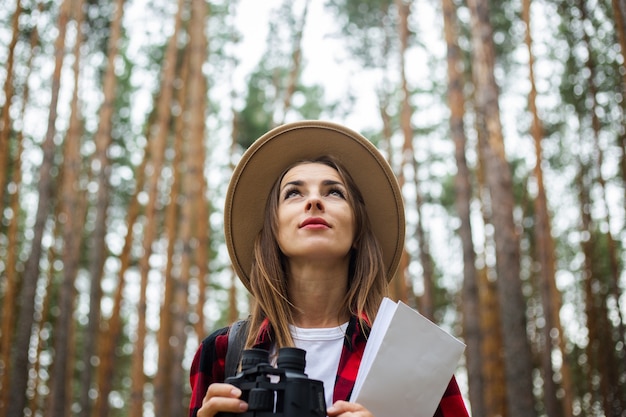 Turista joven en un sombrero, camisa a cuadros roja sostiene un mapa y binoculares en el bosque.