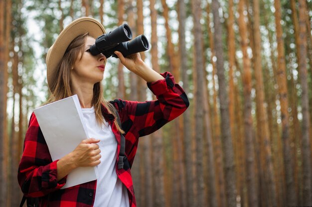 Turista joven con sombrero y camisa a cuadros roja sostiene un mapa y mira a través de binoculares