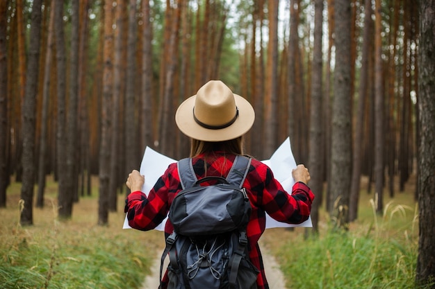 Turista joven en un sombrero, camisa a cuadros roja tiene un mapa de la zona en el bosque.