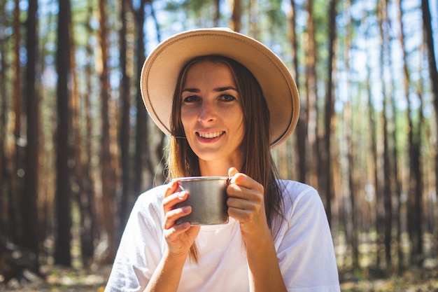 Turista joven con sombrero y camiseta bebe té o agua durante una parada en el bosque.