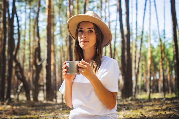 Turista joven con sombrero y camiseta bebe té o agua durante una parada en el bosque.