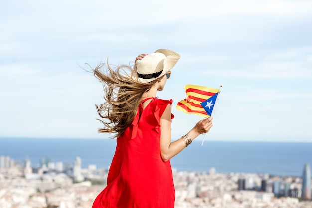 Turista joven en vestido rojo con sombrero y bandera catalana disfrutando de una gran vista del paisaje urbano de Barcelona