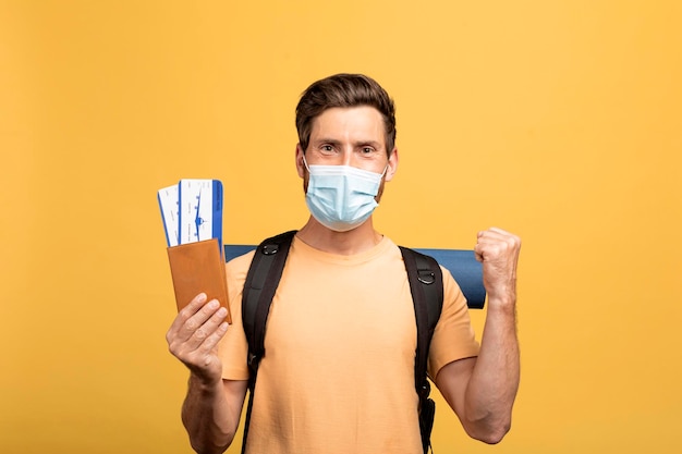 Turista masculino com mochila usando máscara médica segurando bilhetes com passaporte e apertando o punho