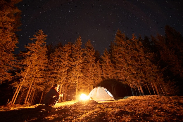 Turista masculino descansar en su campamento cerca del bosque por la noche, sentado cerca de la fogata y la carpa