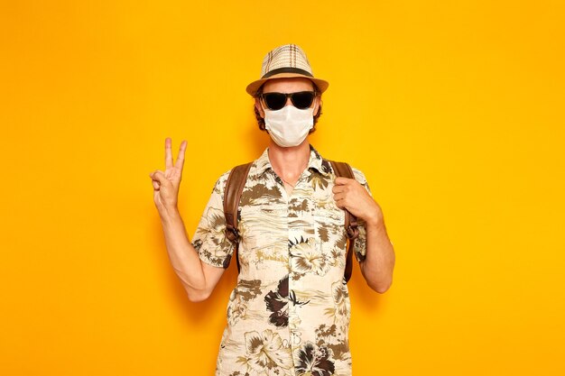 Turista masculino en gafas de sol una máscara médica muestra un signo de paz sobre un fondo amarillo con su mano