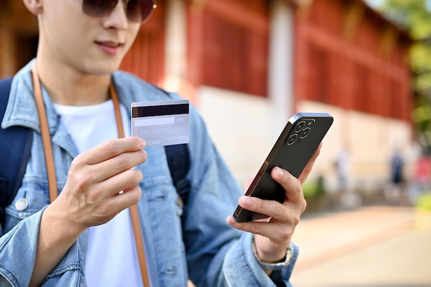 Turista masculino sosteniendo un teléfono inteligente y una tarjeta de crédito usando su aplicación de banca móvil recortada