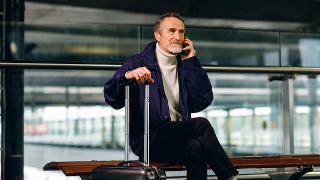 Turista mayor sentado en la terminal del aeropuerto y hablando por teléfono celular