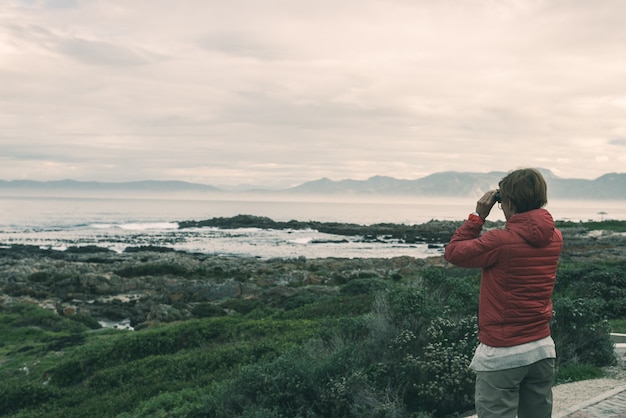 Foto turista mirando con binoculares en la costa rocosa en de kelders, sudáfrica, famosa por la observación de ballenas.