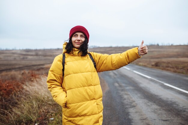 Turista con una mochila con chaqueta amarilla y sombrero rojo coge un coche en la carretera.