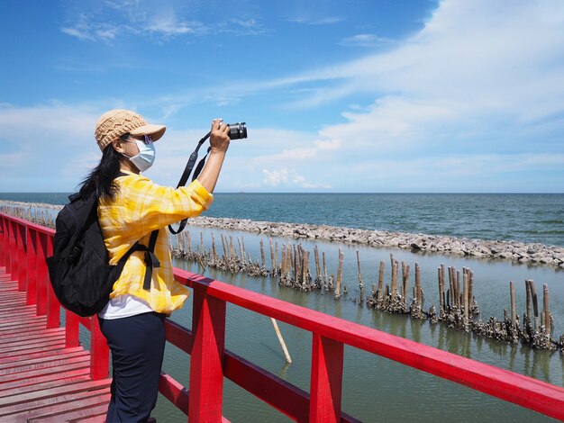 Turista de mujer asiática con mascarilla de pie en el puente rojo y tomar fotografías de la playa de verano en el mirador, provincia de Samutsakhon, Tailandia.