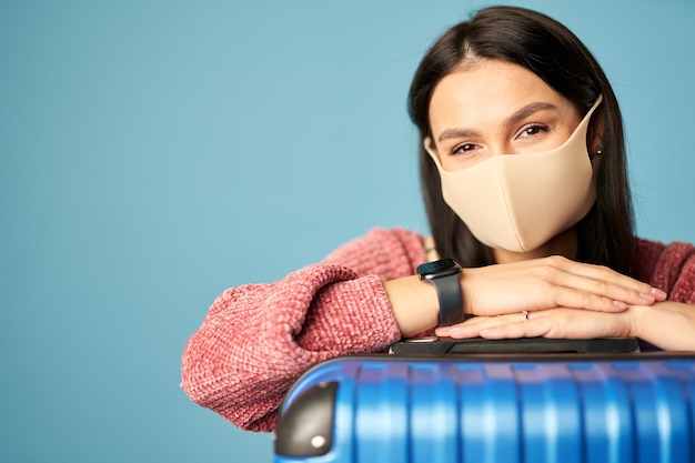 Turista mujer caucásica con una máscara antivirus con maleta posando sobre fondo azul. Copie el espacio. Concepto de viaje, coronavirus.